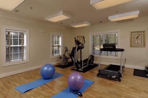 A fitness room with wooden flooring featuring two blue exercise mats each with a large exercise ball on top. In the background, there are three pieces of cardio equipment including a stationary bike, an elliptical machine, and a treadmill. The room has three windows with white blinds, beige walls, and ceiling lights. A framed picture hangs on the wall to the right.