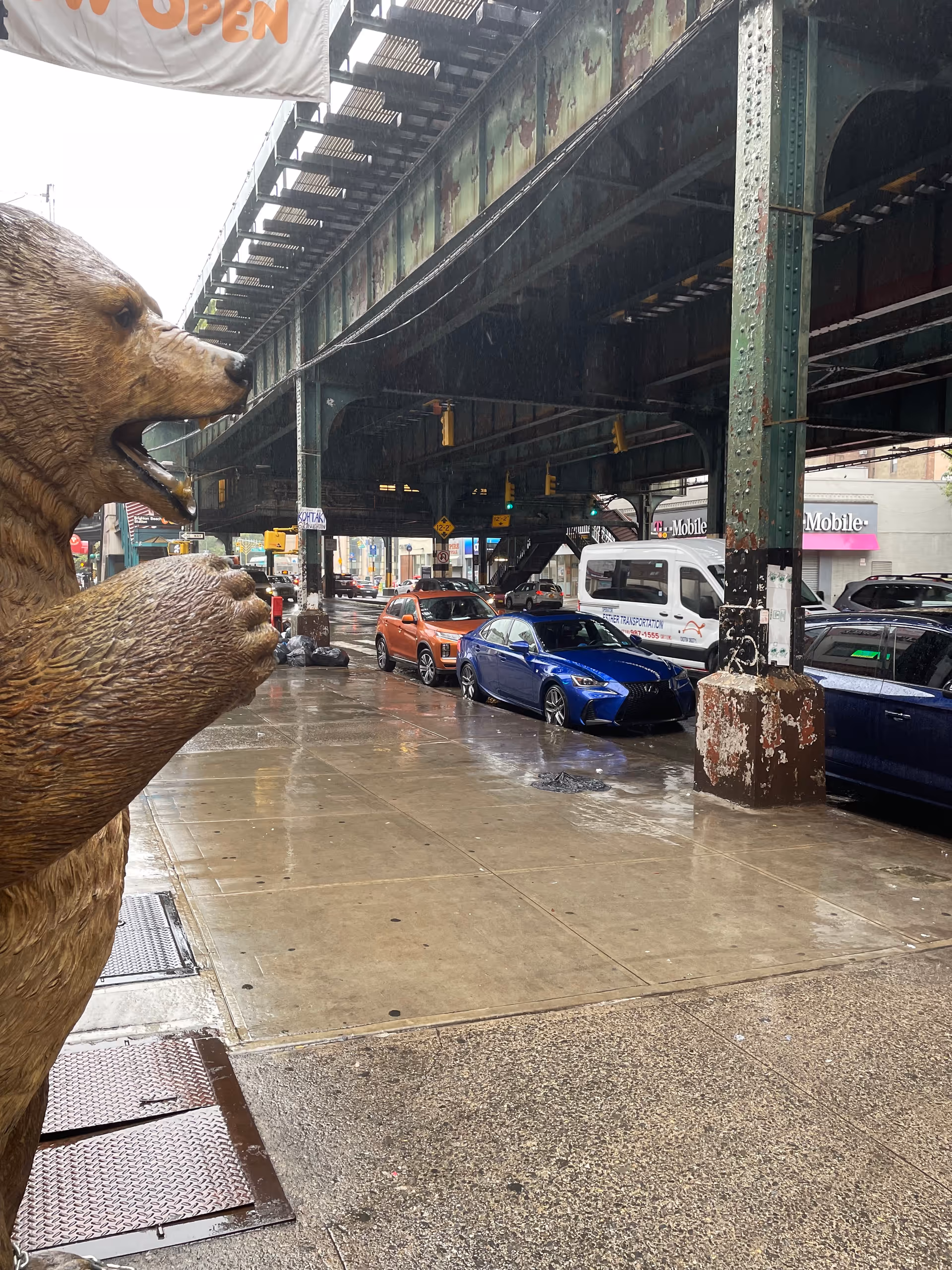 Rainy city street under elevated tracks with a large bear statue in foreground.