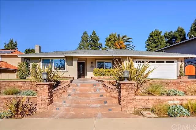 Single-story ranch-style home with front steps, brick planters, landscaping, and an attached garage under a clear blue sky.