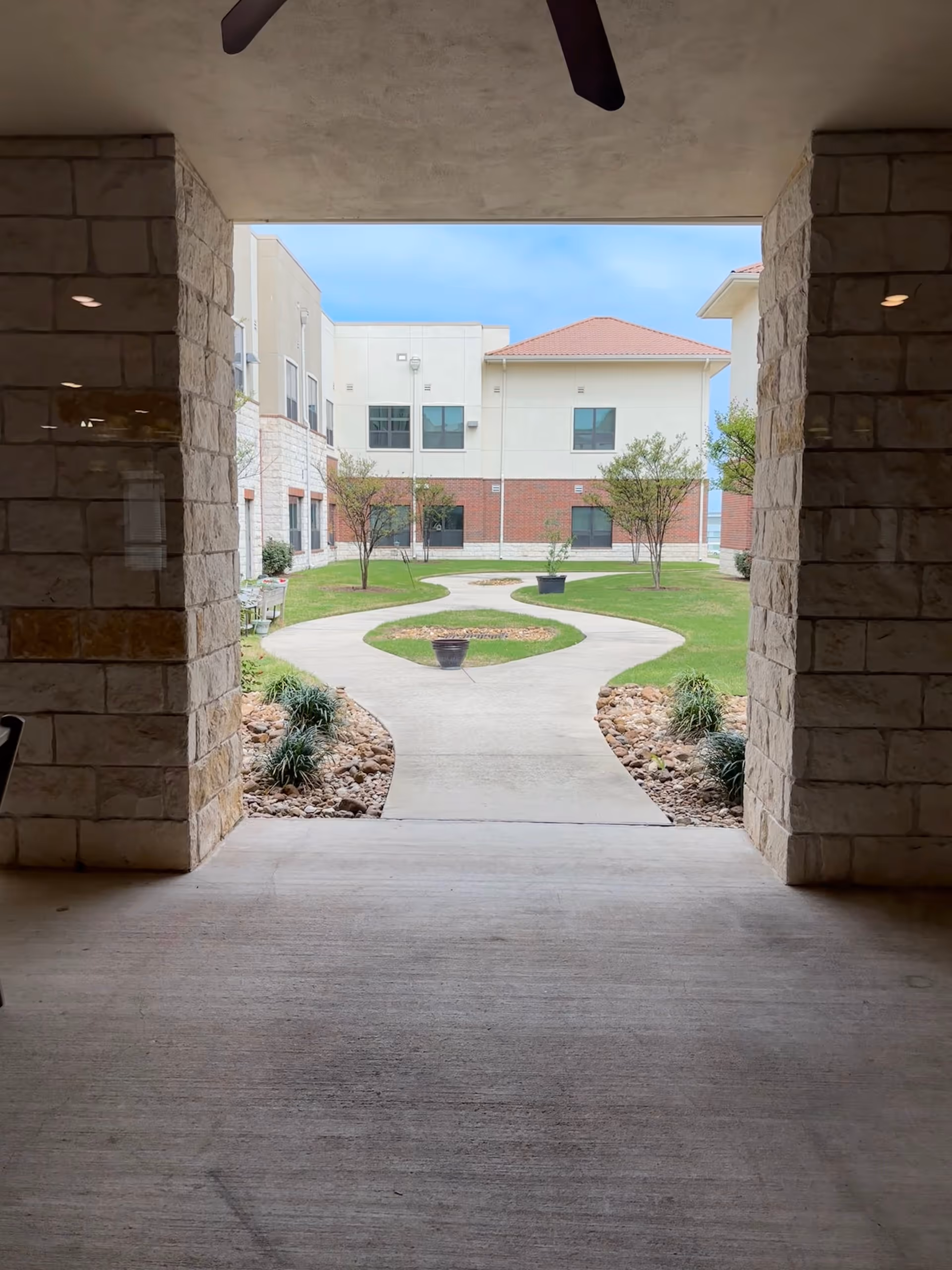Covered walkway frames a sunny courtyard with curved paths, small trees, and adjacent residential building.