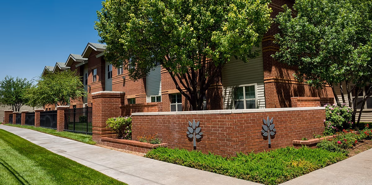 Exterior view of a brick senior living facility building with multiple windows, surrounded by green trees, bushes, and a well-maintained lawn. A sidewalk runs alongside the building under a clear blue sky.