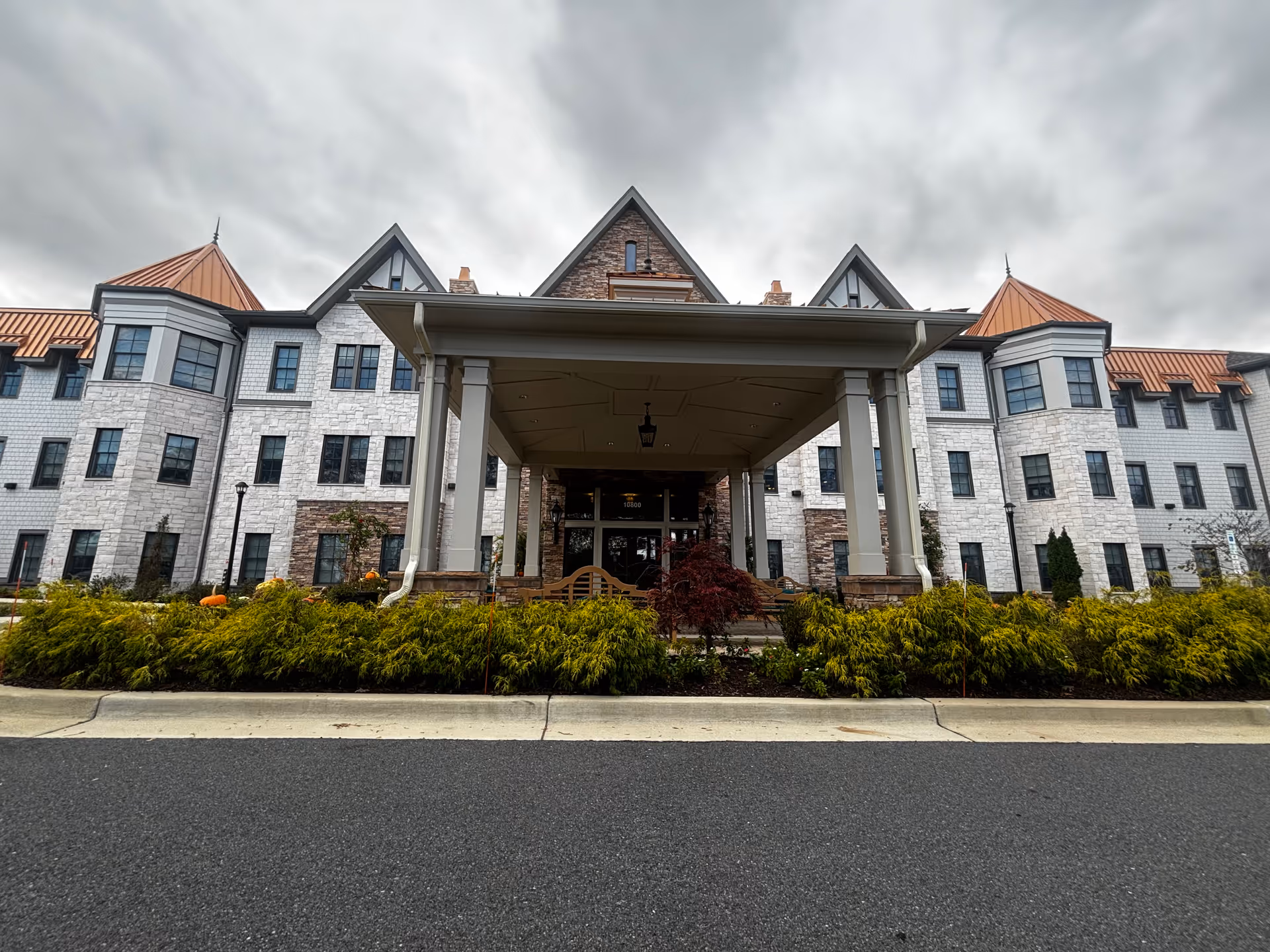 Front entrance of a multi-story senior living building with a covered porte-cochère, stone facade, and landscaped shrubs under a cloudy sky.