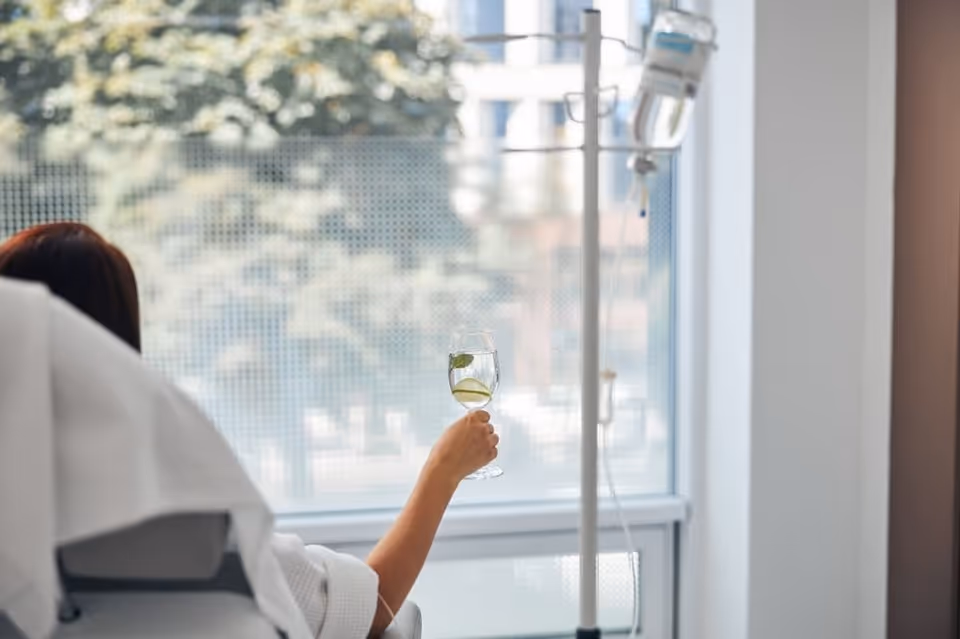 Patient in a hospital-style chair holds a glass beside an IV pole.