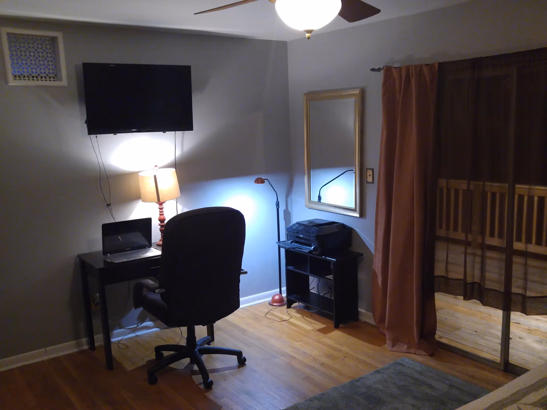 Compact living room workspace with desk, laptop, chair, lamp, printer, wall TV, mirror, and brown curtains.