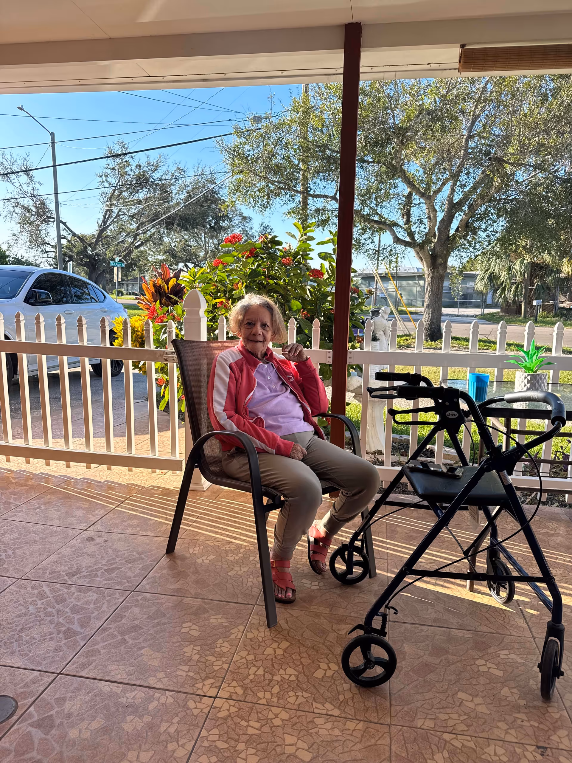 Elderly woman sits on a chair on a covered outdoor porch with a walker nearby.