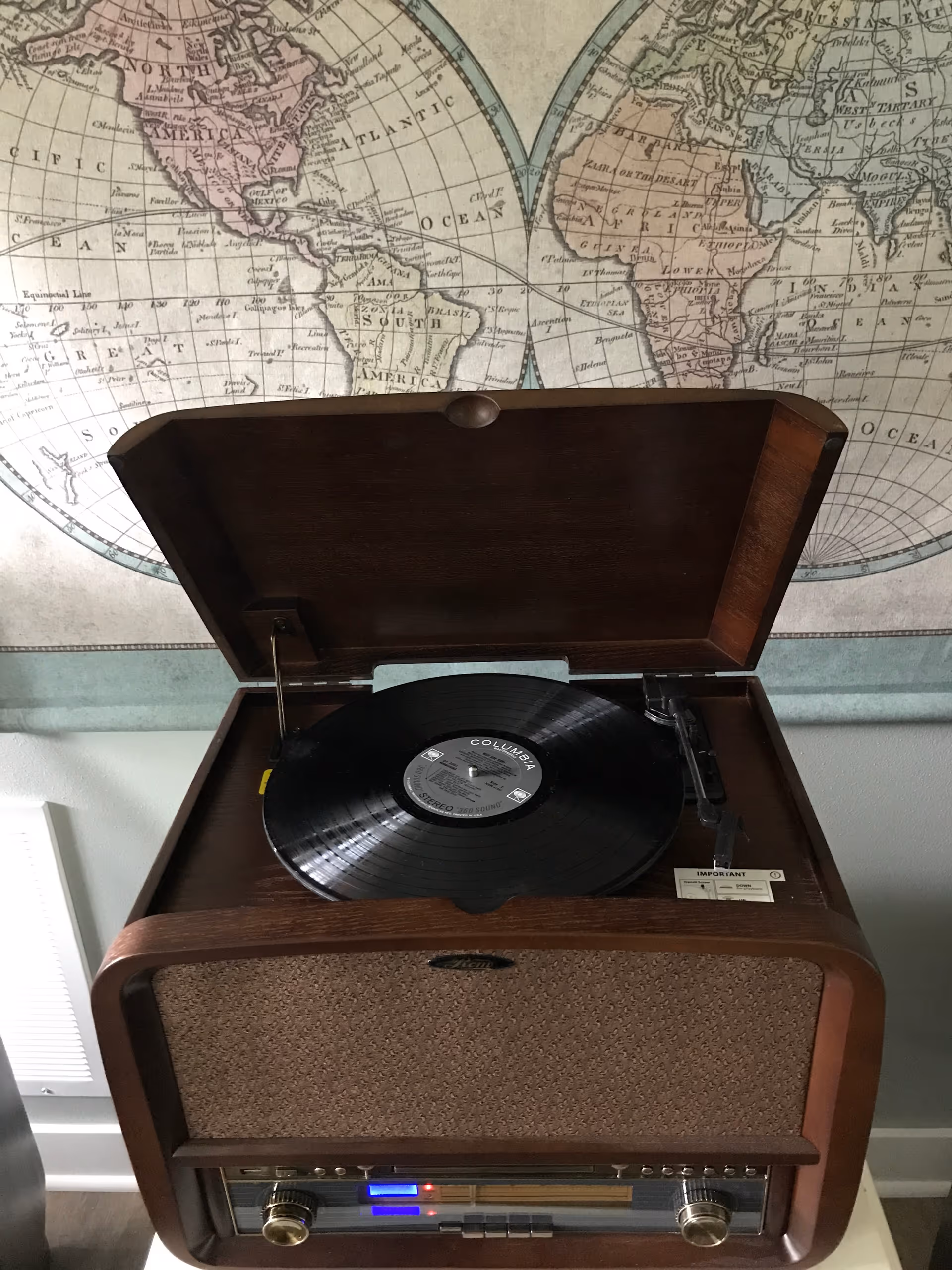 A vintage-style record player with a vinyl record on the turntable, set against a wall with a large world map. The record player has a wooden exterior and a fabric speaker cover, with control knobs and a display panel below.