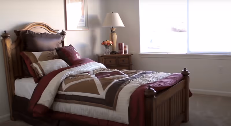 A sunlit bedroom with a wooden bed dressed in brown and burgundy bedding, a nightstand with a lamp and decorative items, and a large window.