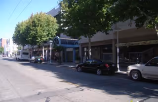 Exterior street view of a building with storefronts, trees, and parked cars.