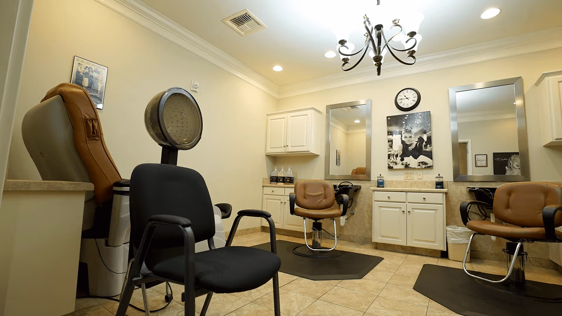 Small salon area with multiple styling chairs, mirrors, a hooded hair dryer, cabinetry and a chandelier.