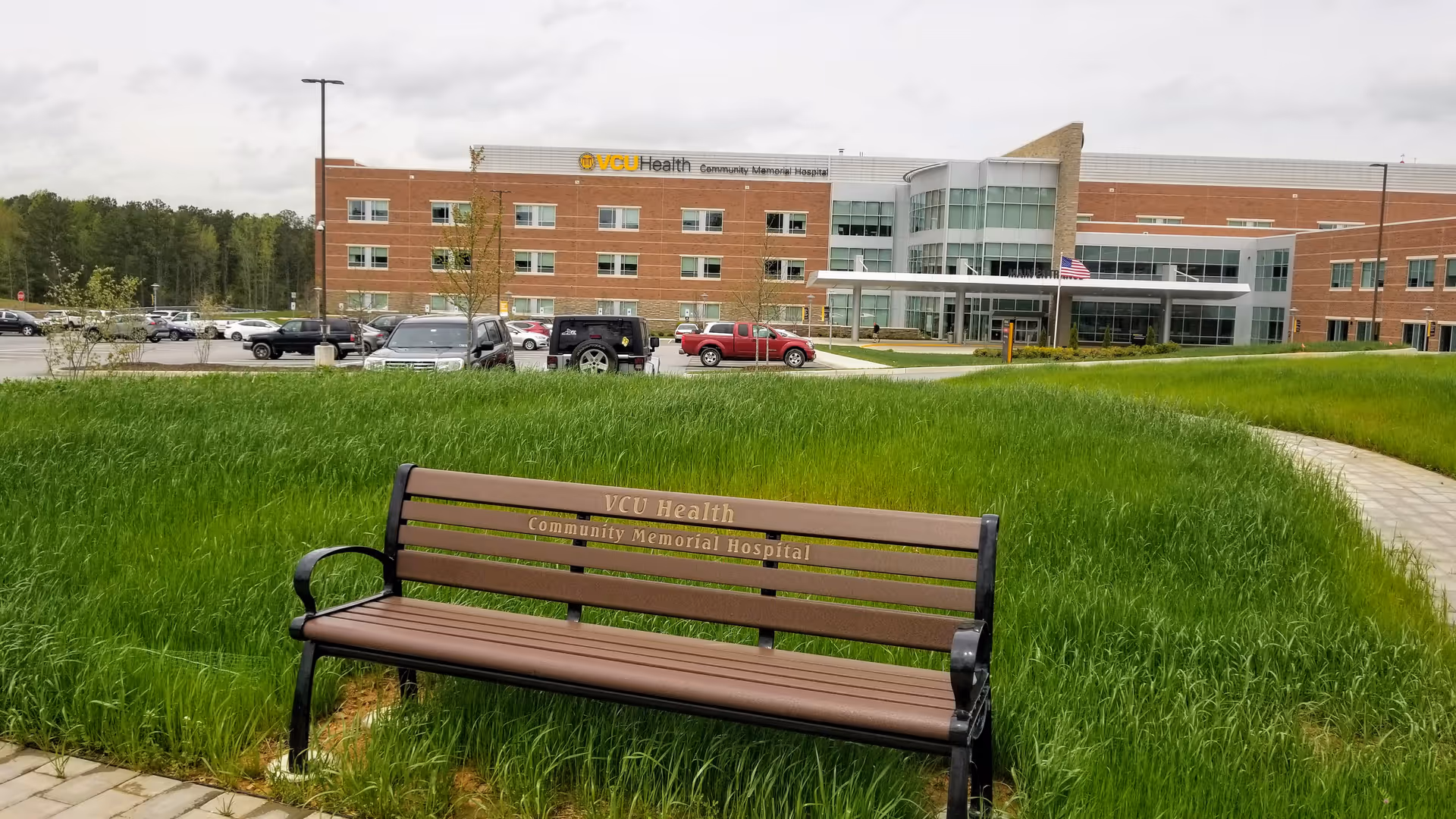 A bench sits on a grassy area with the hospital building in the background.
