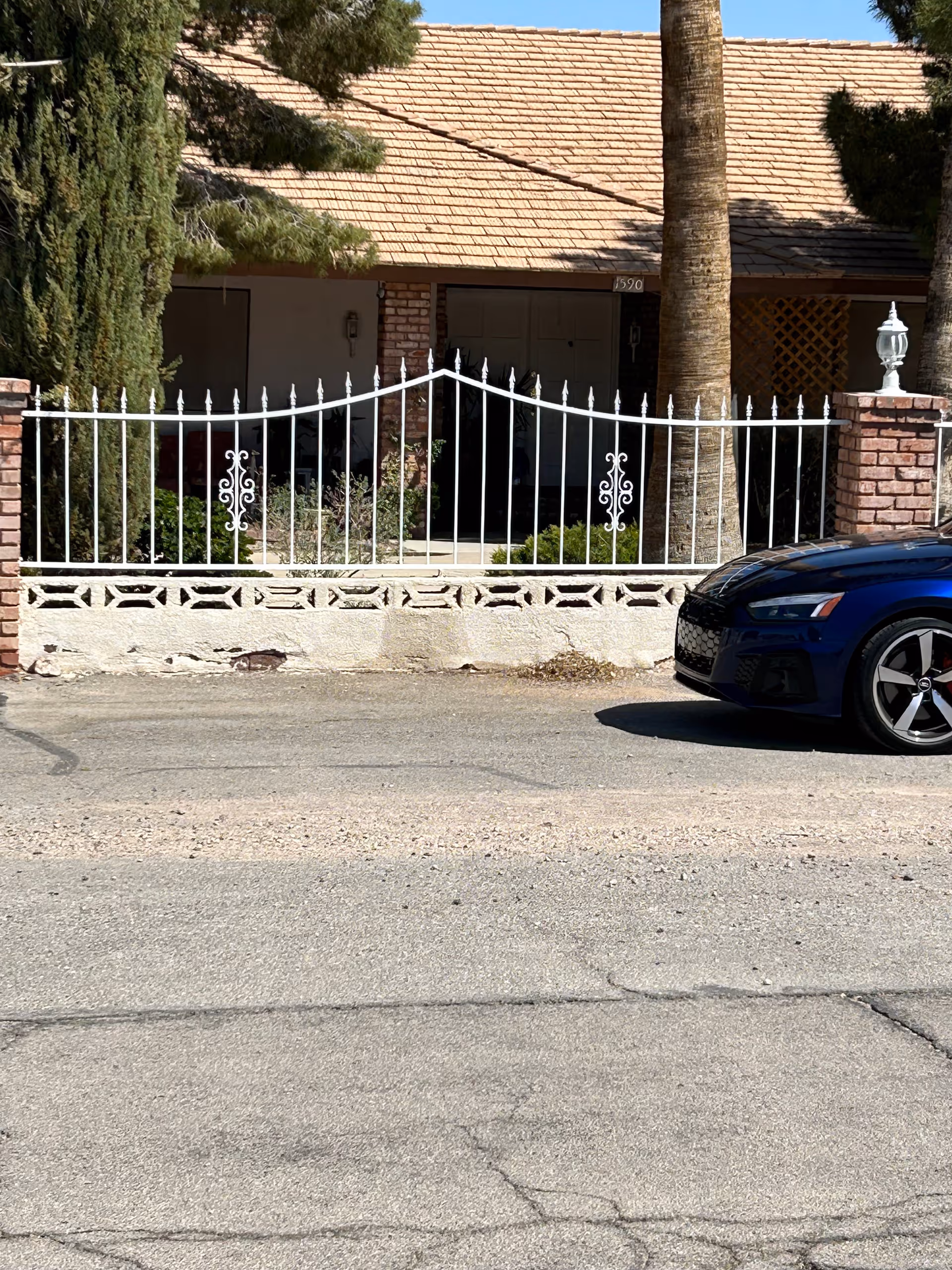 Front exterior view of a single-story house with a tiled roof, brick pillars, a white metal fence, and a blue car parked on the street in front. There are trees and shrubs around the house.