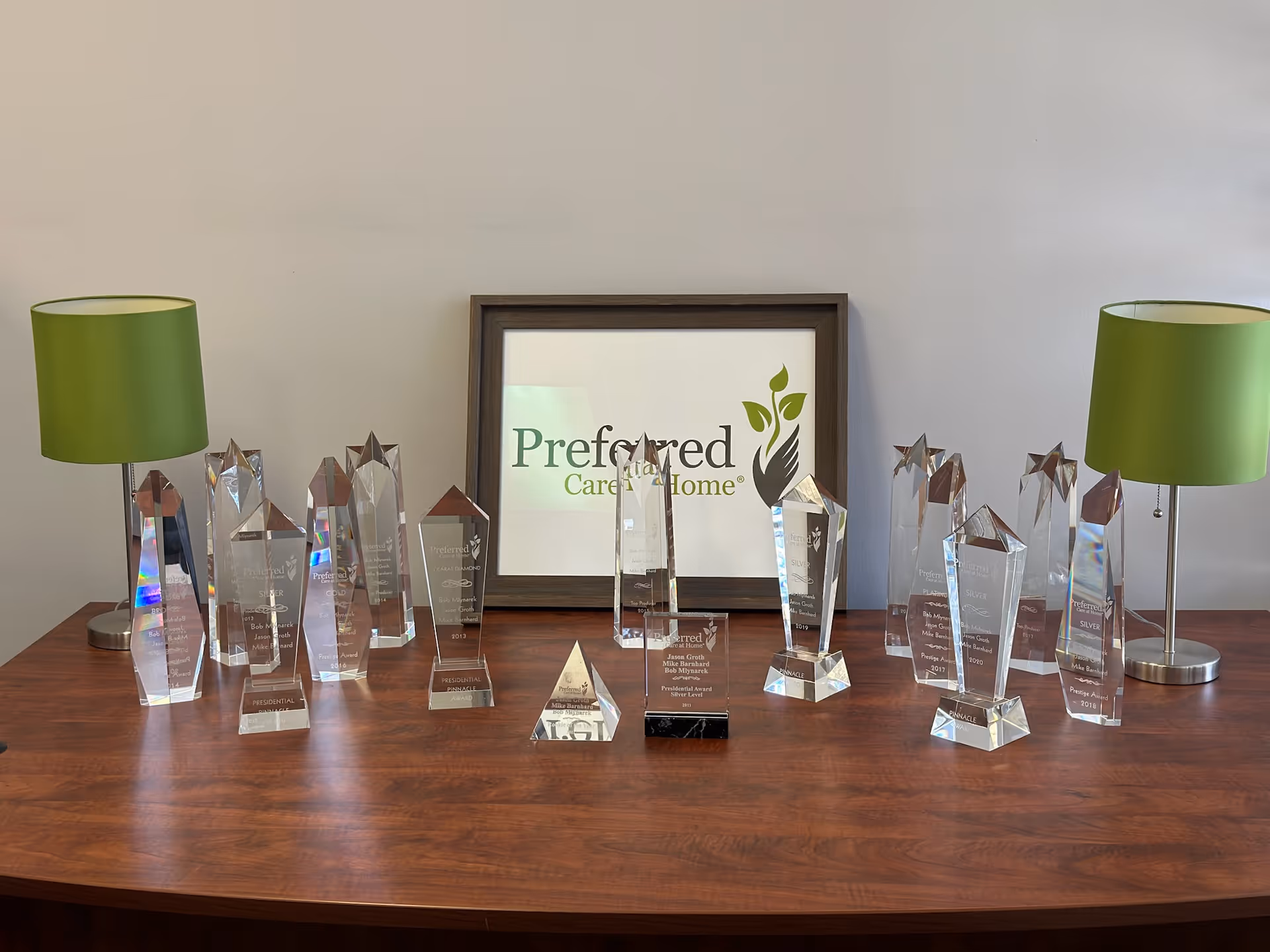 A wooden table displays numerous glass trophies with a framed sign reading Preferred Care at Home.