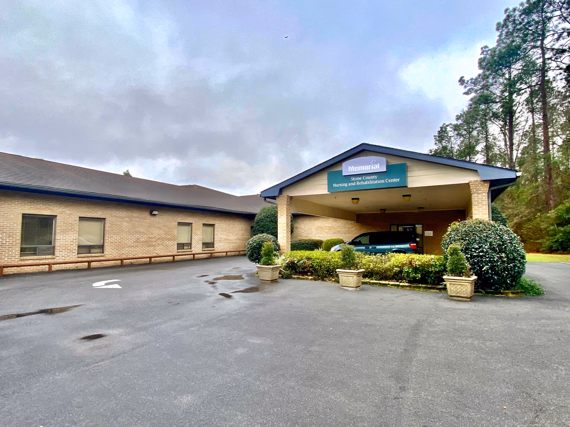 Exterior view of Stone County Nursing and Rehabilitation Center showing a single-story brick building with a covered entrance. There are shrubs and potted plants around the entrance, a parked vehicle under the canopy, and trees in the background under a cloudy sky.