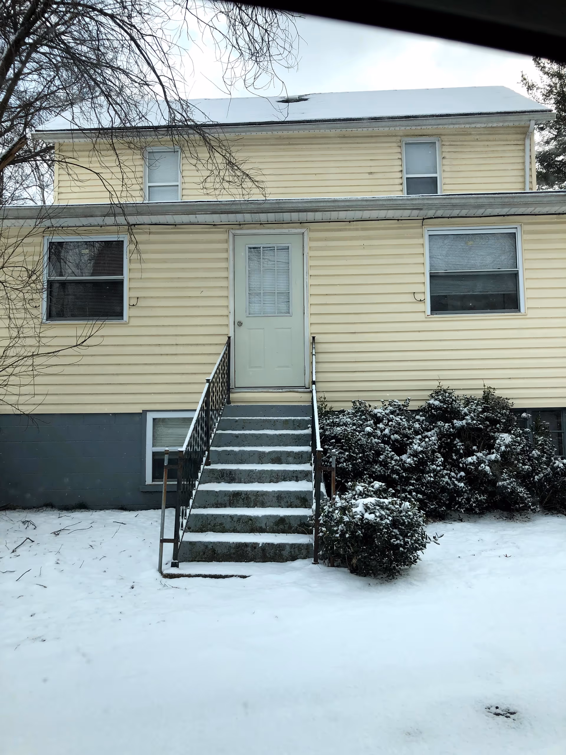 Two-story yellow building with a front entry door, stairs, and snow-covered ground.