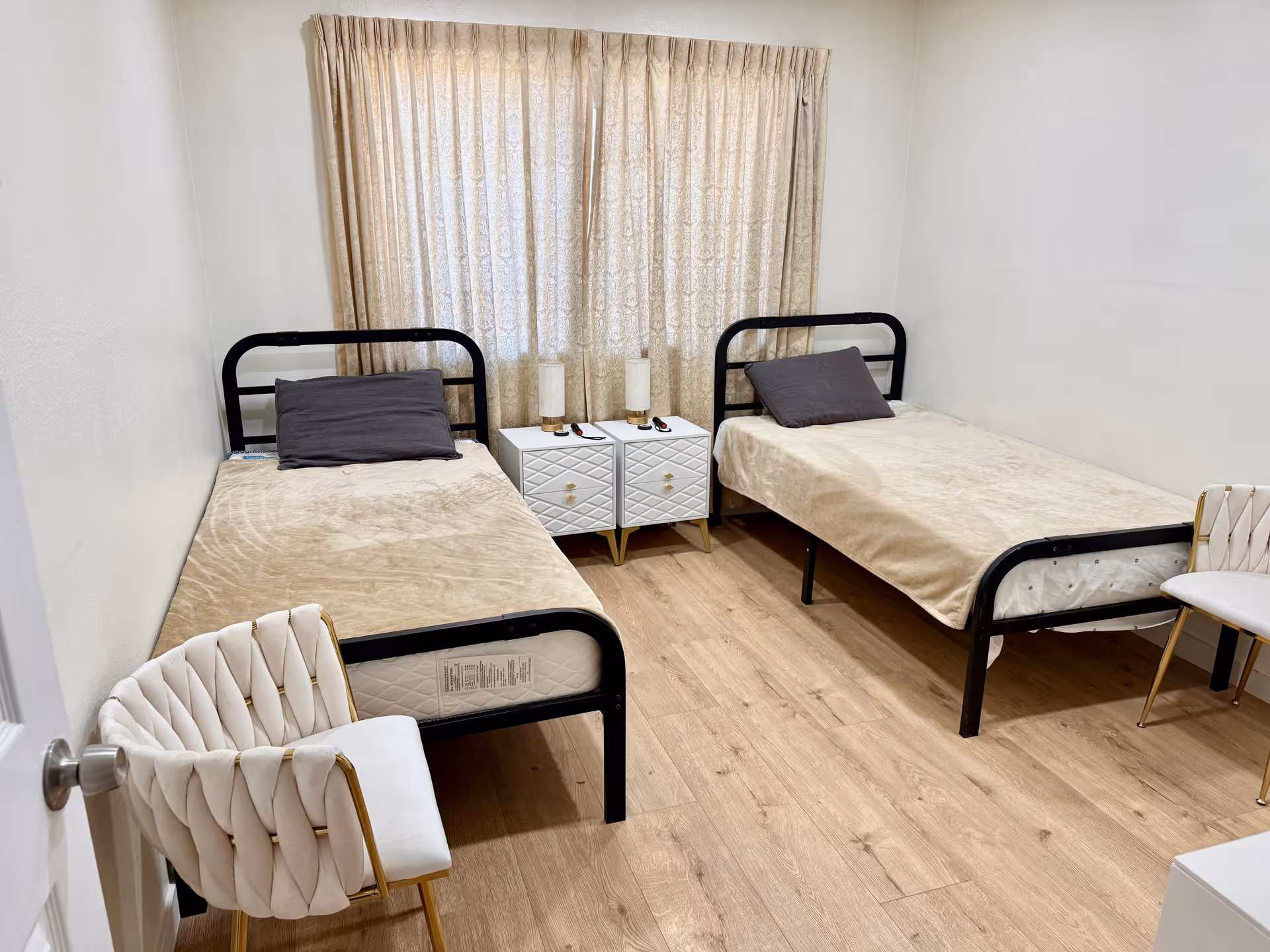 Two single beds with black frames, beige bedding, nightstands, lamps, and chairs in a small bedroom.