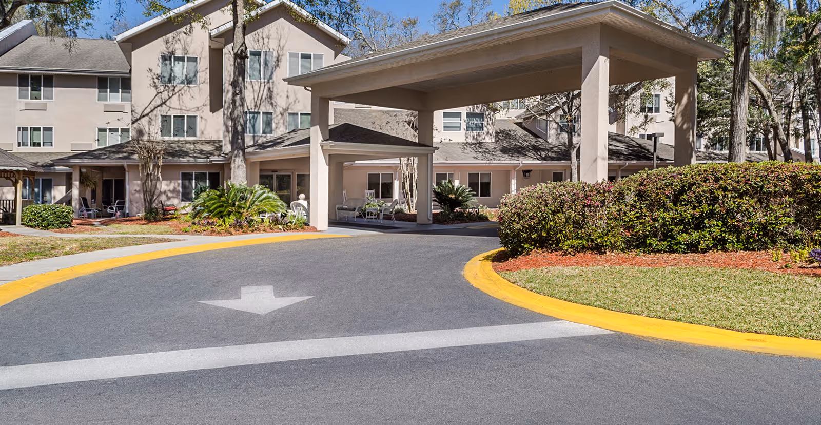 Exterior view of Holiday Indigo Pines senior living facility showing a multi-story building with a covered entrance driveway, landscaped bushes, and trees under a clear blue sky.