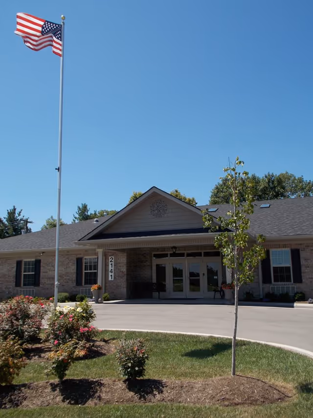 Front exterior view of a single-story brick building with a peaked roof, an American flag on a tall flagpole, a small tree, and landscaped flower beds in front. The building has large glass double doors and windows with black shutters.