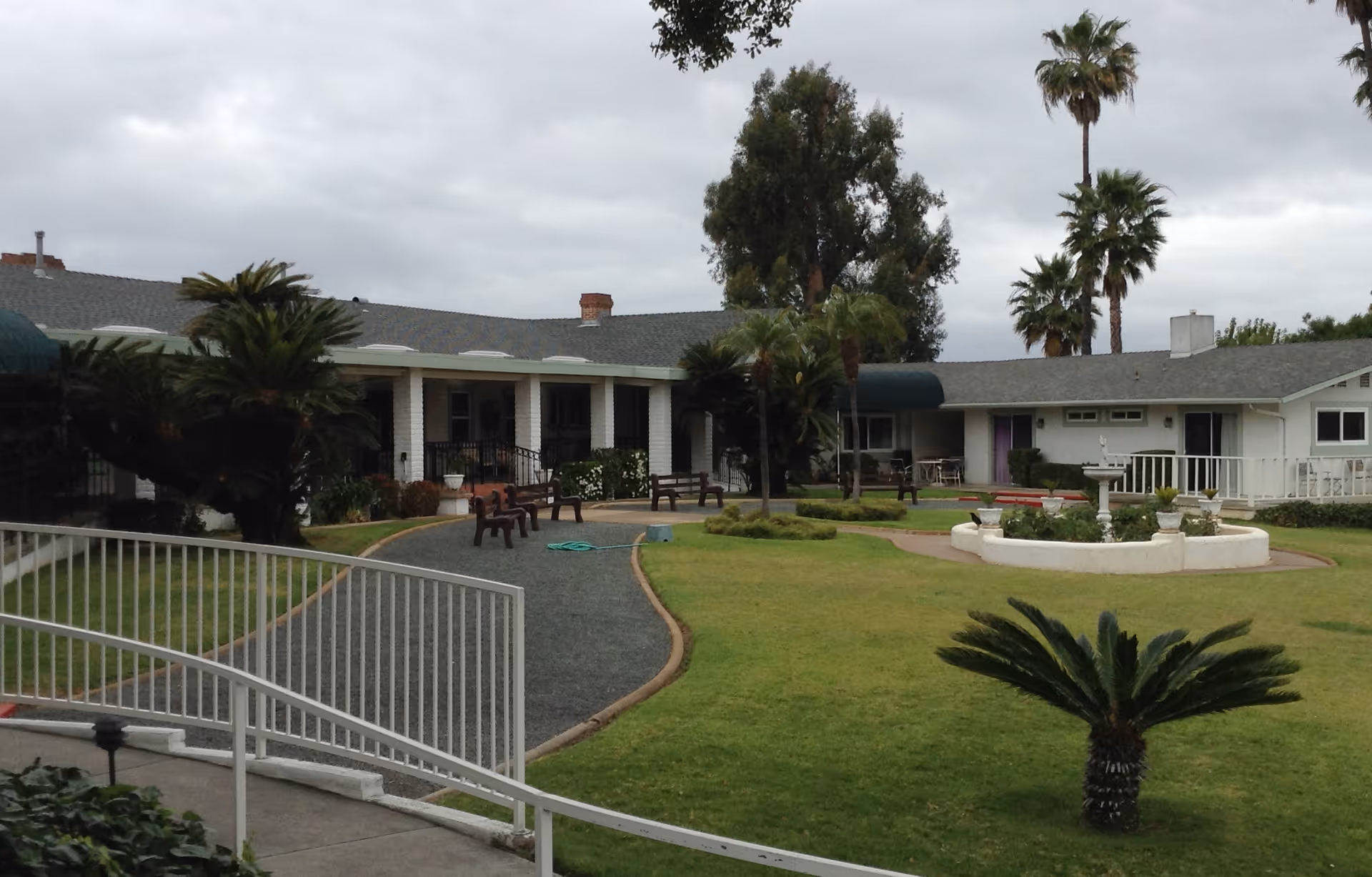 Outdoor courtyard area of Crestview Manor featuring a green lawn, palm trees, a white circular planter with a birdbath, several benches along a curved gravel path, and a single-story building with a gray roof and white exterior walls in the background under a cloudy sky.