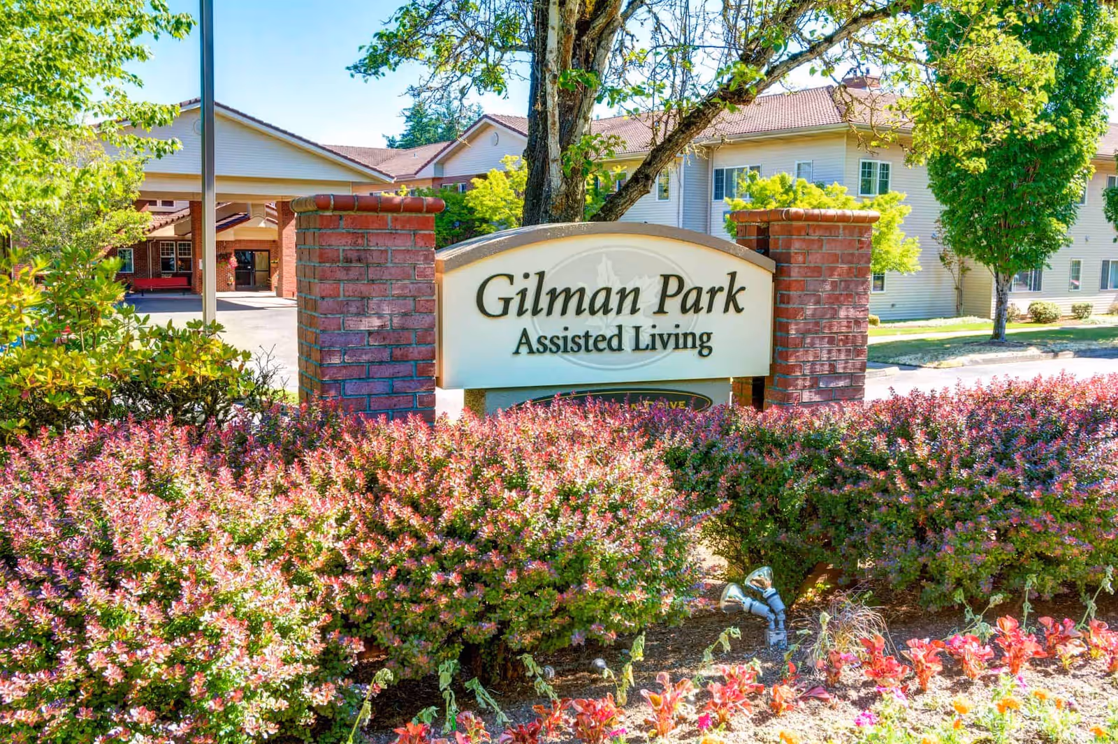 Entrance sign reading 'Gilman Park Assisted Living' surrounded by landscaping with the facility building in the background.