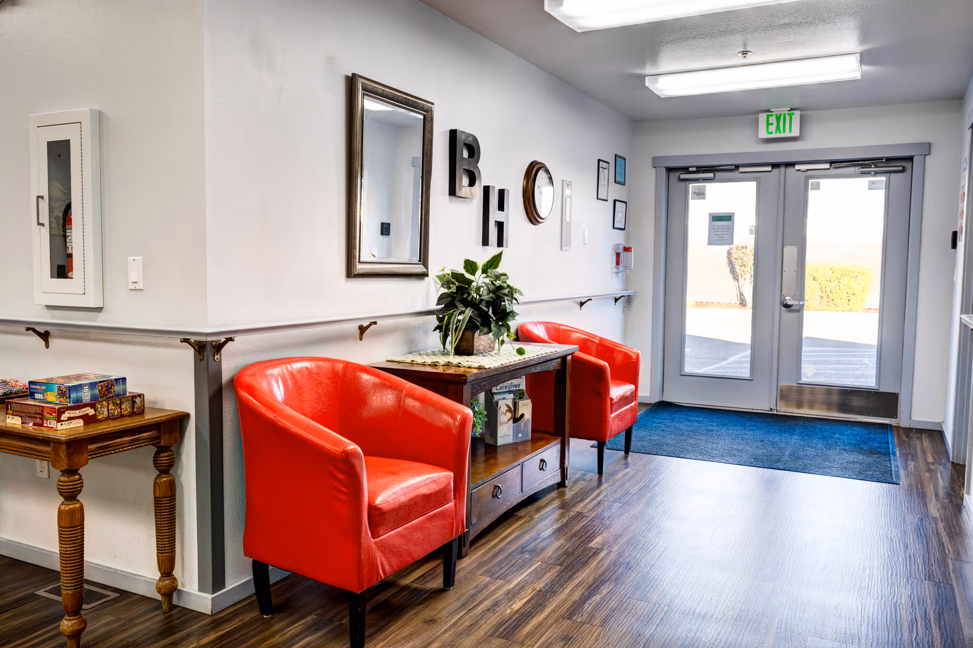 A bright hallway with two red armchairs placed on either side of a wooden table with a plant and books. There is a mirror and decorative letters on the wall above the table. A small wooden side table with board games is on the left. Double glass doors with an exit sign lead outside. The floor is wood with a blue mat near the doors.