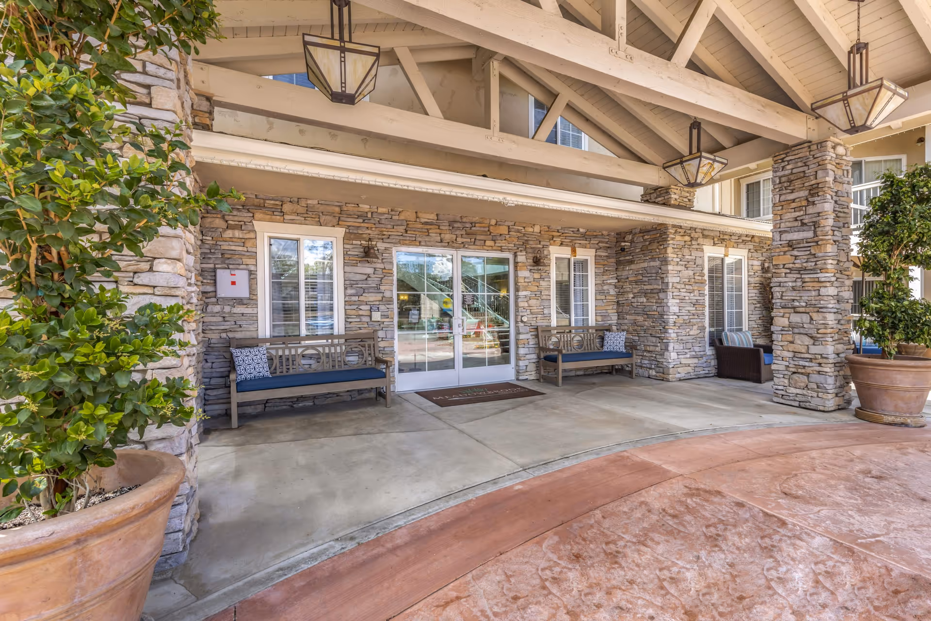 Covered stone-faced entrance with benches, large potted plants, and glass double doors under a wooden beam canopy.