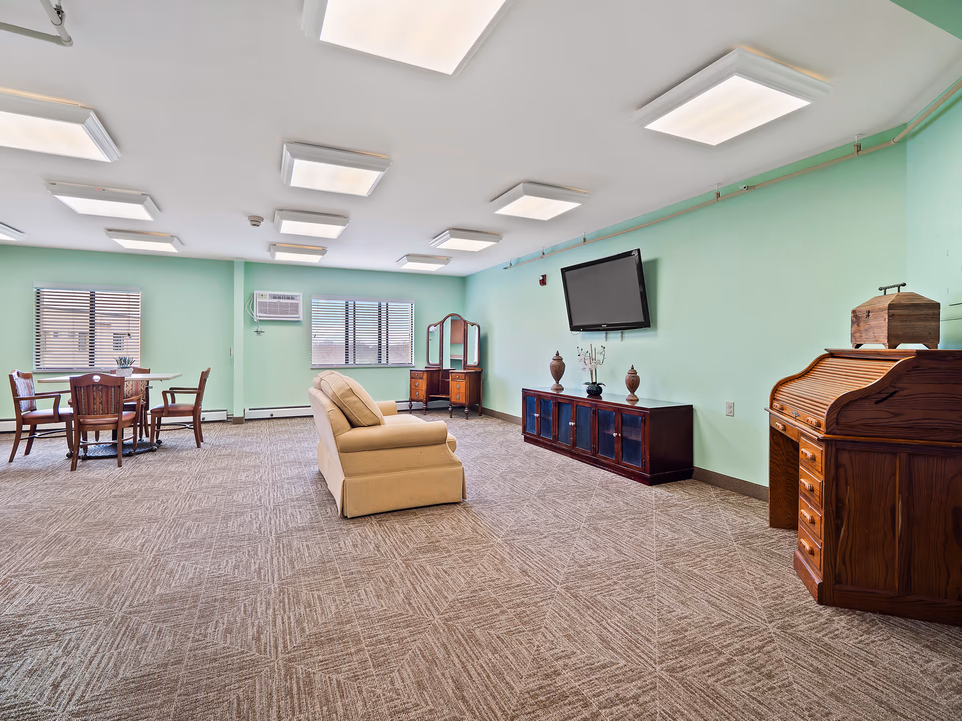 Spacious senior living lounge with mint walls, seating, a TV, and a dining area.
