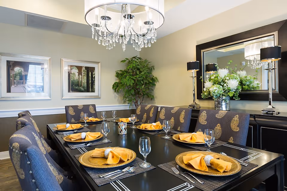 A formal dining room with a long black dining table set for eight people. Each place setting includes a gold charger plate, a folded yellow napkin with a napkin ring, a fork, knife, spoon, and a clear water glass. The chairs are upholstered with a dark fabric featuring a gold paisley pattern. On the table, there is a small decorative candle holder. The room has beige walls, two framed pictures, a large mirror, two table lamps with black shades, a chandelier with hanging crystals, and a large green plant in the corner.