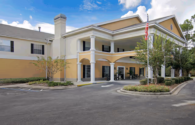 Front entrance of a two-story beige senior living building with columns, a covered porte-cochère, and an American flag.