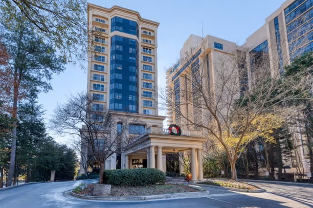 Exterior view of a tall senior living facility building with a covered entrance, surrounded by trees and landscaping under a clear blue sky.