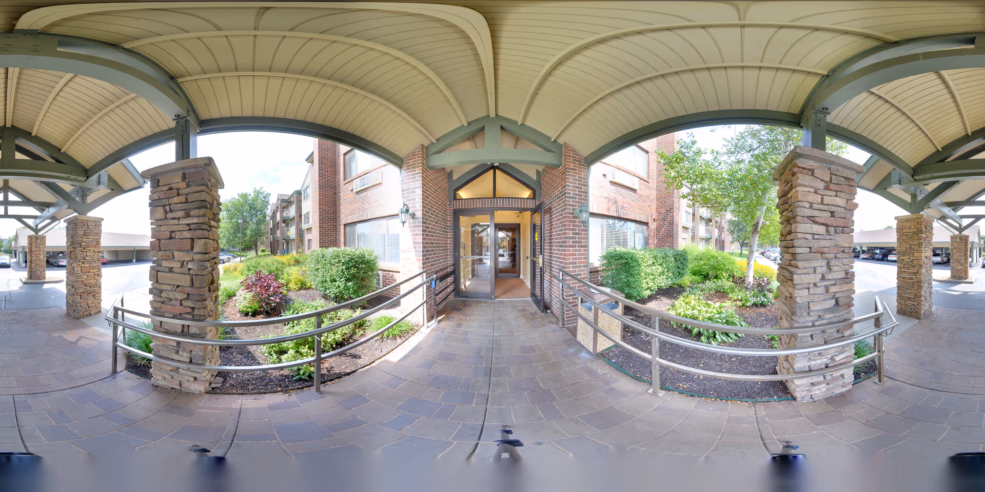 Covered entrance to a brick building with stone pillars and metal railings, surrounded by landscaped greenery and a paved walkway.
