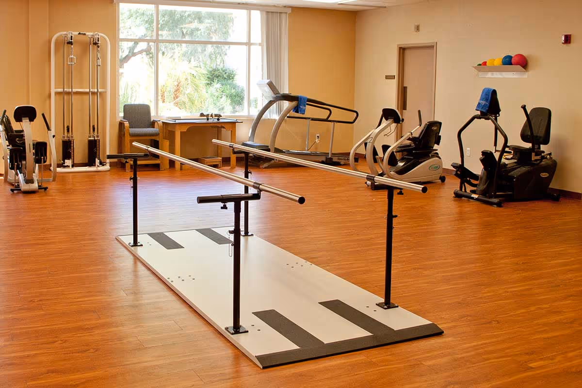 A bright rehabilitation/exercise room with parallel walking bars, treadmills, stationary bikes, and other fitness equipment on a wood floor.