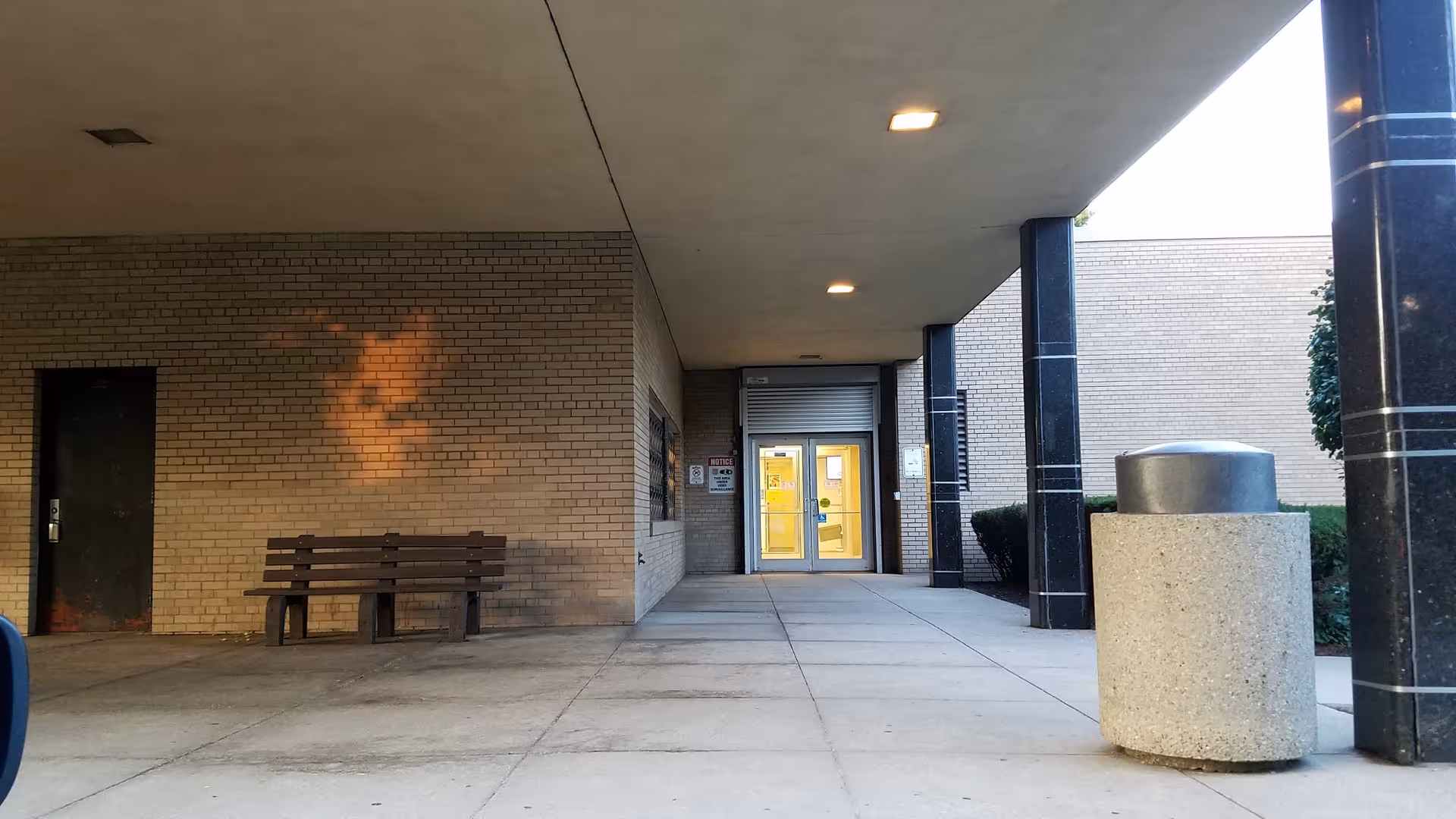 Covered walkway to brick building entrance with a bench and glass doors.