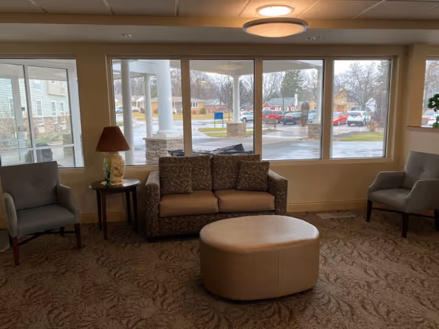 Seating area with a sofa, two armchairs, round ottoman and table lamp in front of large windows overlooking the facility entrance and parking lot.
