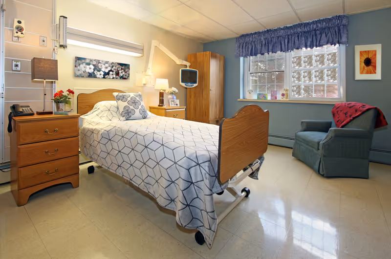 A well-lit patient room in a healthcare facility featuring a single hospital bed with a geometric patterned blanket and a decorative pillow. The room includes wooden furniture such as a nightstand with a lamp, a phone, and flowers, a small cabinet with a lamp and framed photo, and a wardrobe. There is a blue armchair with a red blanket near a window with blue curtains. Medical equipment is mounted on the wall near the bed.