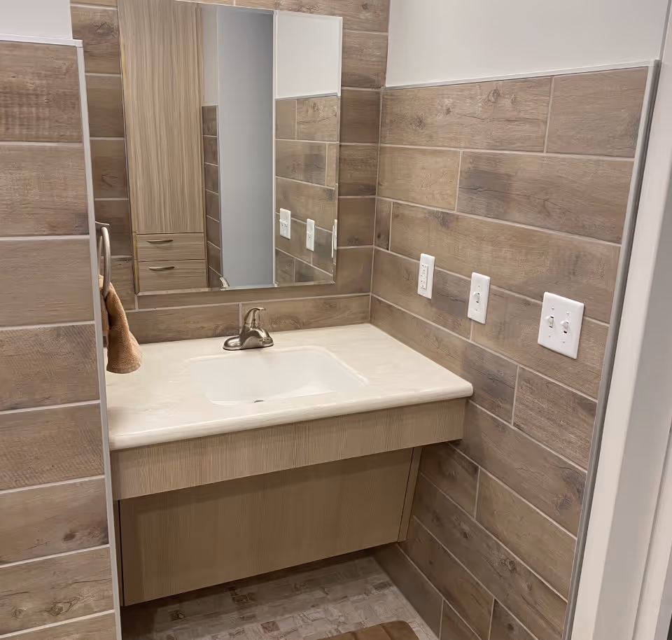 A bathroom vanity with sink, mirror, and wood-toned tile walls in a senior facility.