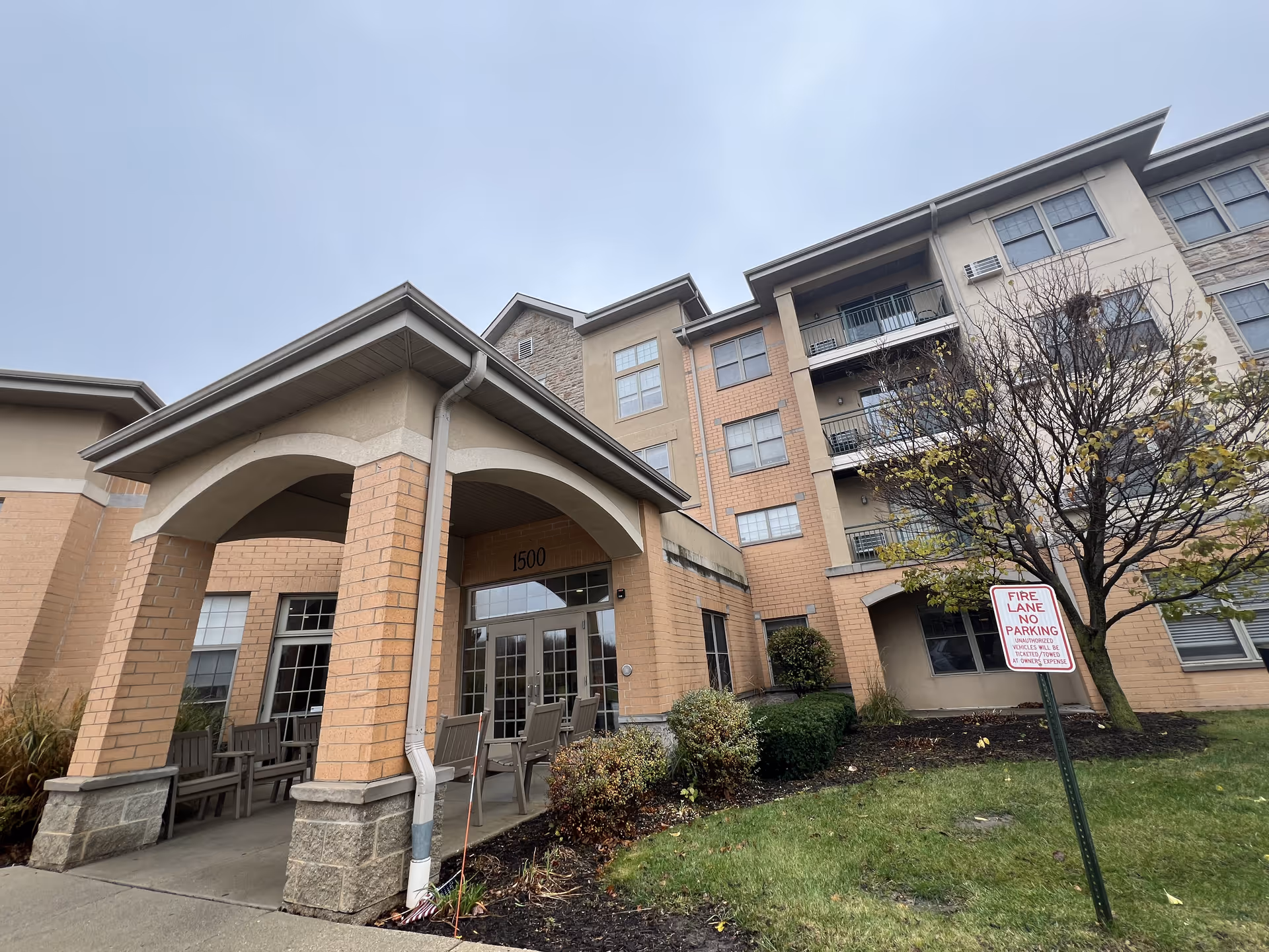 Exterior view of a multi-story assisted living facility building with a covered entrance supported by brick columns. There are chairs arranged under the entrance canopy, some bushes and a tree with sparse leaves in front, and a fire lane no parking sign on the grass. The sky is overcast.