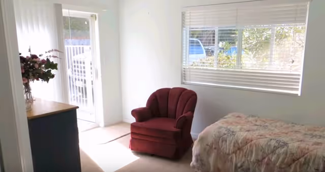 Sunlit bedroom with a bed, red armchair, dresser, flowers, and a window view.