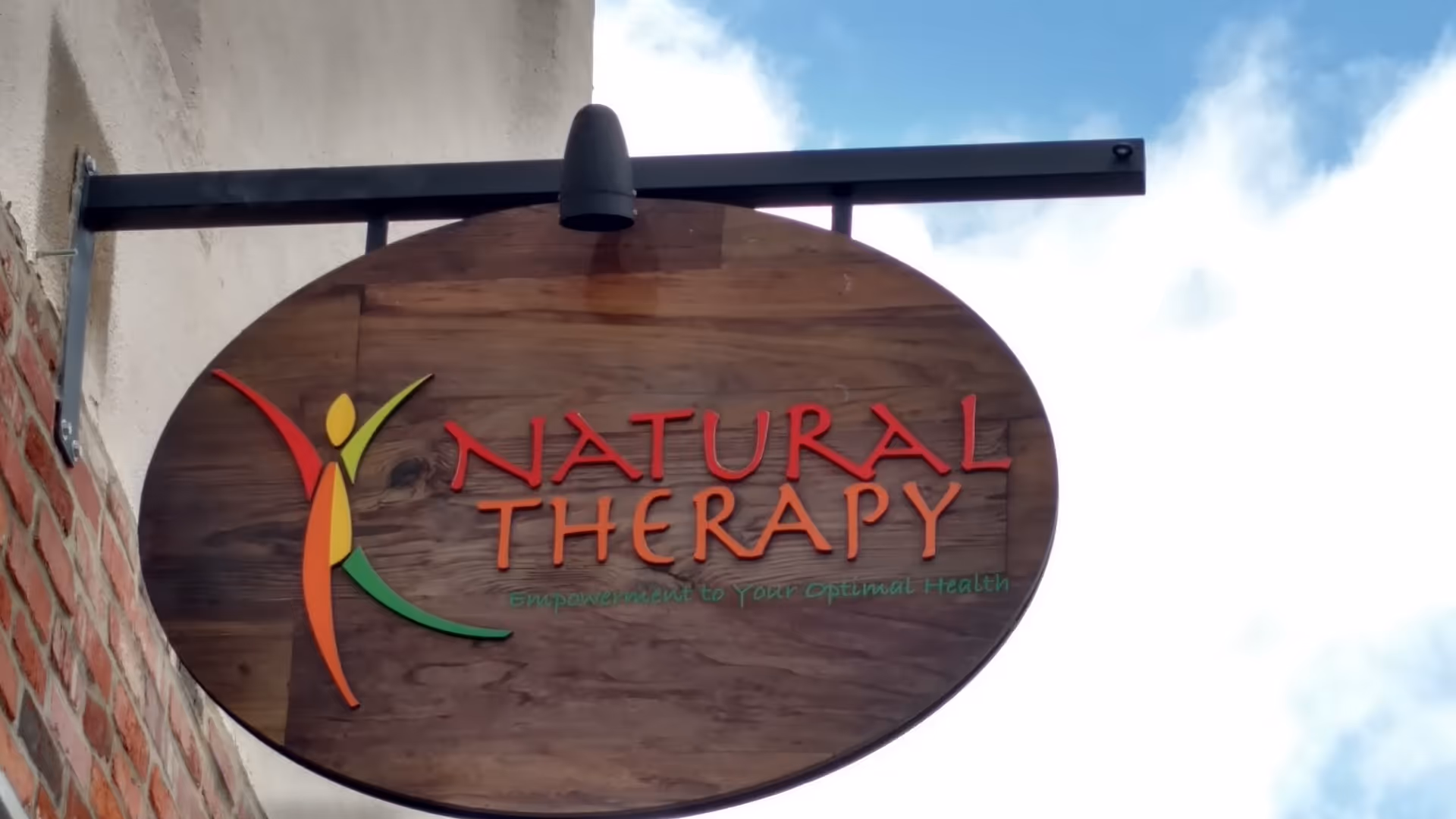 An oval wooden sign reading Natural Therapy with a colorful abstract figure on a brick exterior.