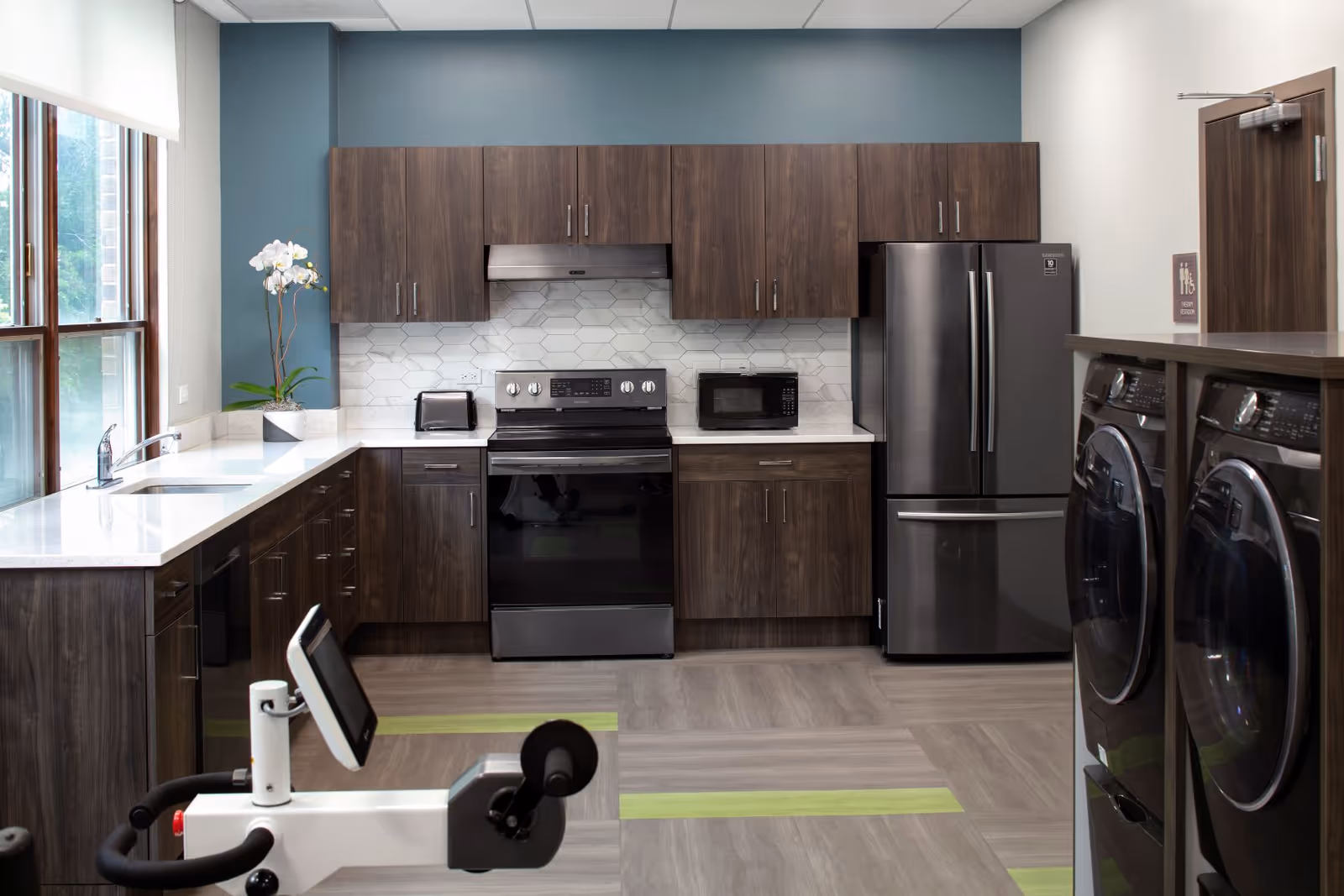Modern communal kitchen with dark wood cabinets, stainless appliances, and a fitness bike in foreground.