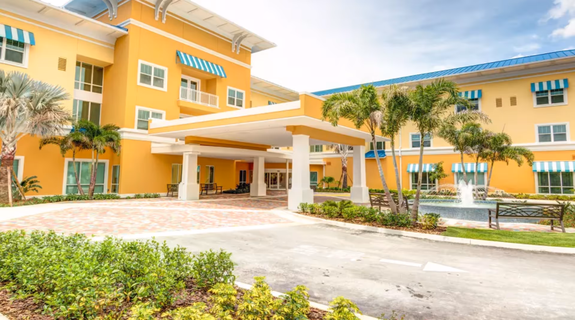 Sunny yellow senior living building with a covered entrance, palm trees, and a fountain courtyard.