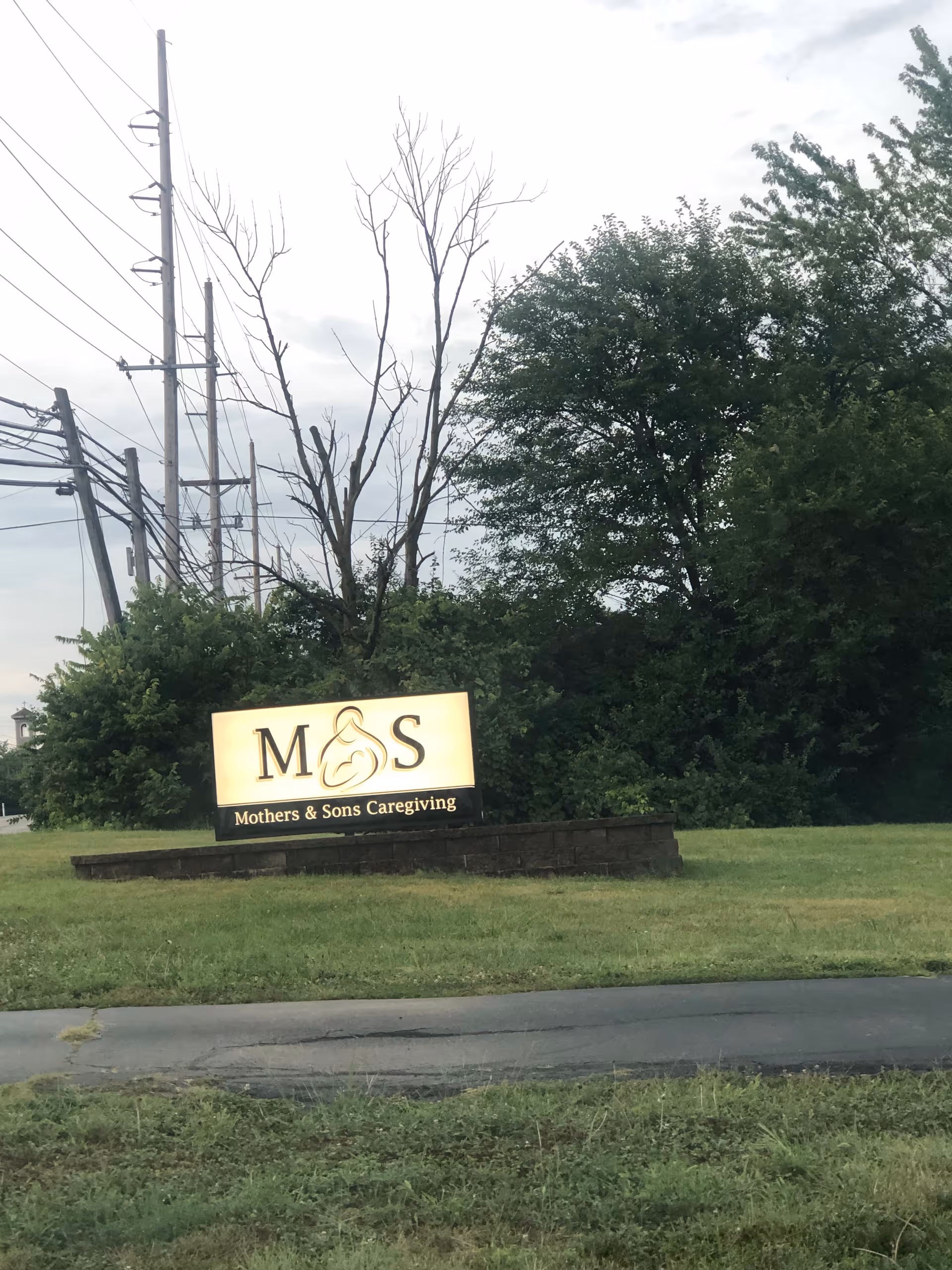 Outdoor sign reads Mothers & Sons Caregiving on a grassy roadside with trees.
