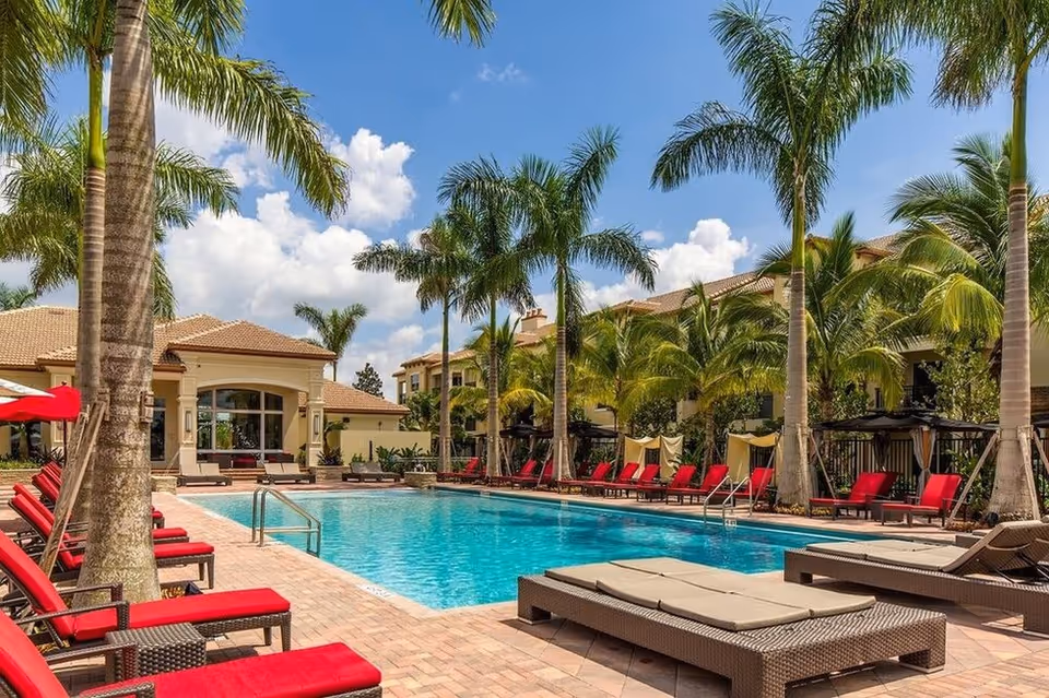 Outdoor swimming pool area at UpsideHoM Pembroke Pines with red cushioned lounge chairs lined up along the poolside, surrounded by tall palm trees and a building with a tiled roof in the background under a partly cloudy blue sky.