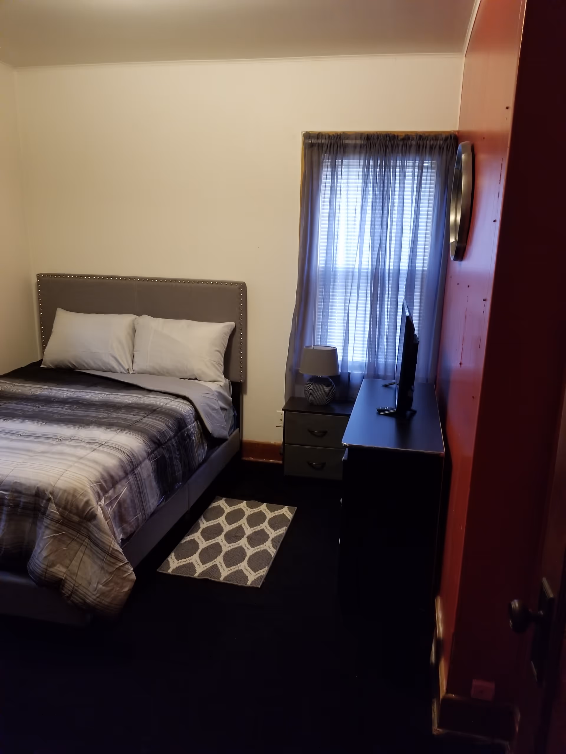 Cozy bedroom with a bed, nightstand, dresser, TV, and window with curtains.