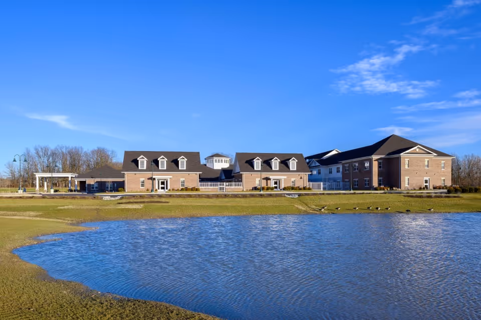 A brick senior living campus building with dormer windows sits behind a grassy lawn and a pond under a clear blue sky.