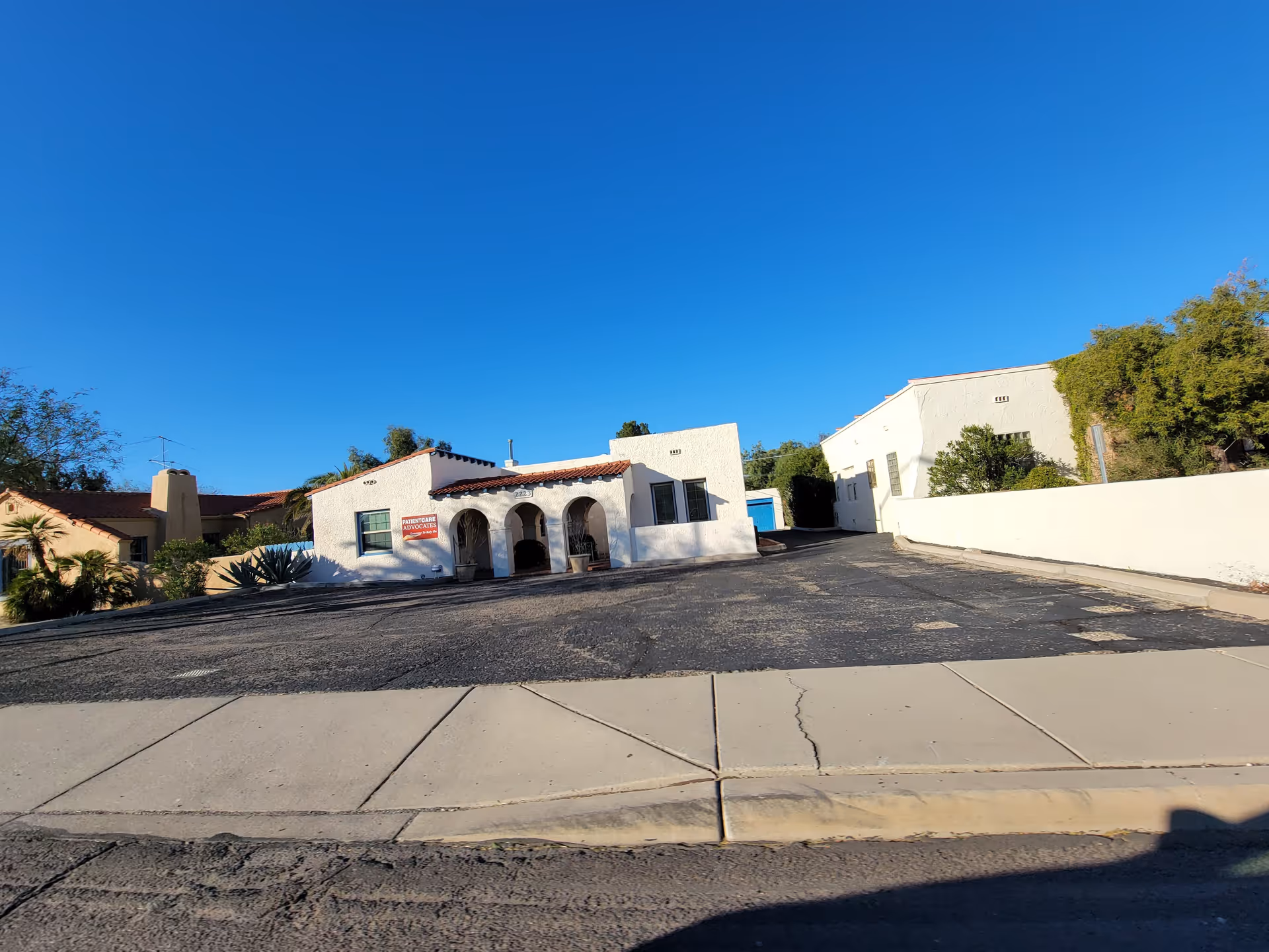 White stucco facility with arched entrances and a broad parking area under a clear blue sky.