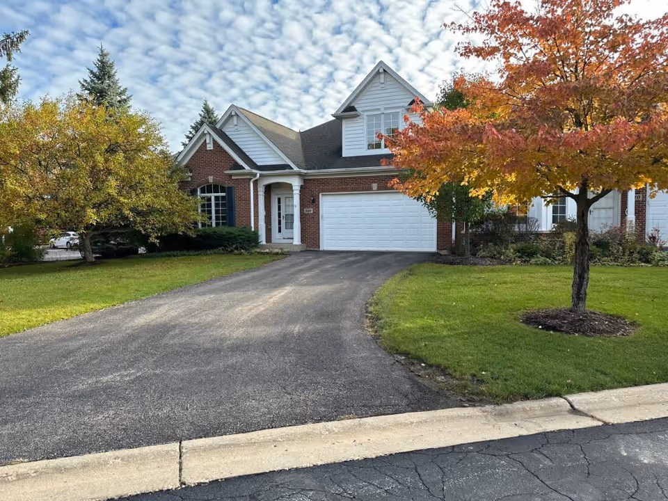 A two-story brick and white siding house with a double garage and a driveway. The house is surrounded by green grass and trees, one of which has autumn-colored leaves. The sky is partly cloudy with a pattern of scattered clouds.