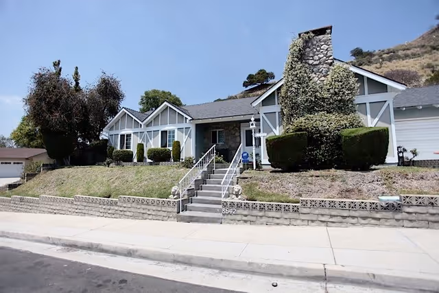 Front view of a single-story house with a stone chimney, steps up to a porch, and trimmed shrubs on a sloped yard.