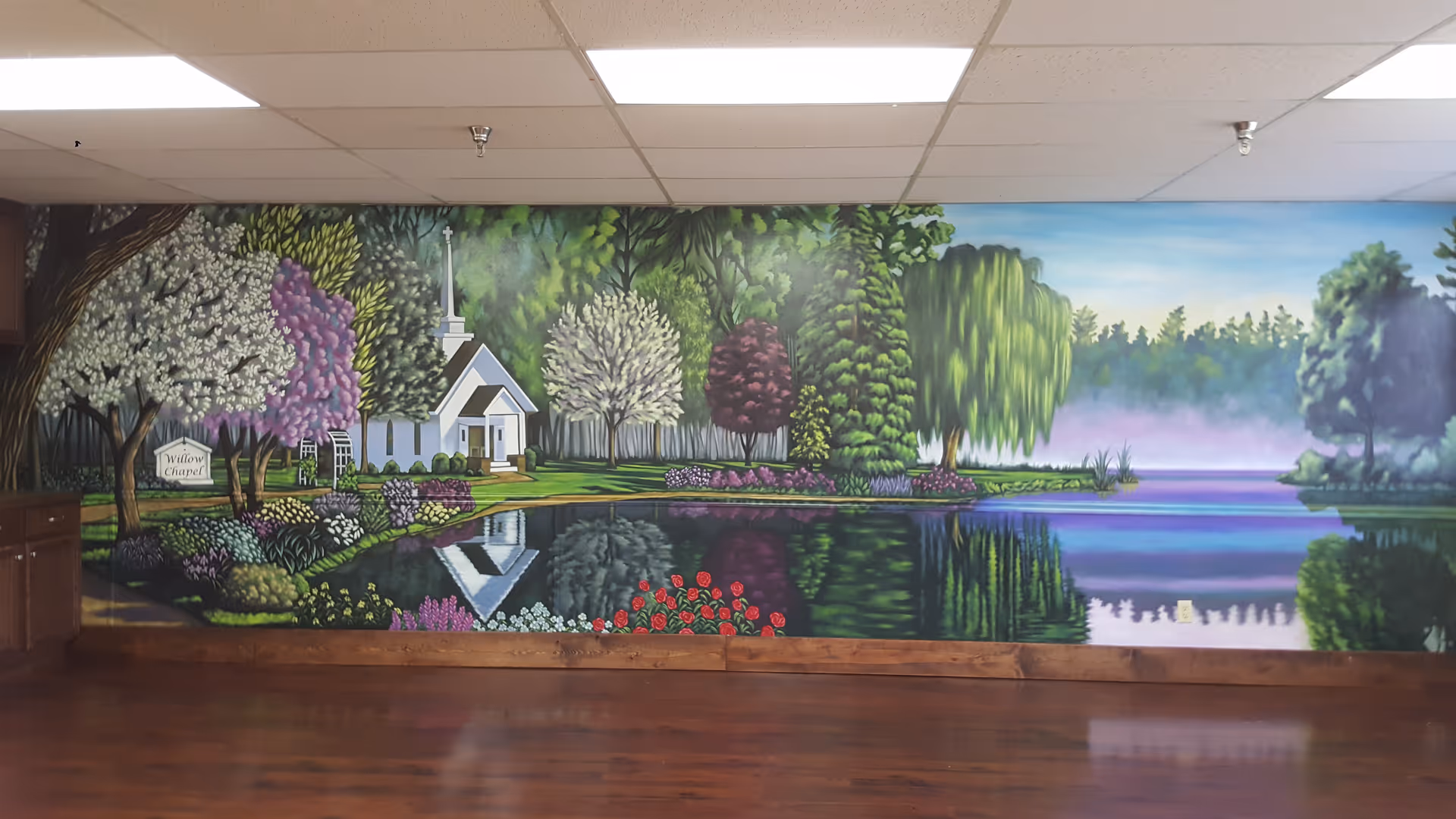 Colorful lakeside mural with trees, flowers, and a small church on an interior wall.
