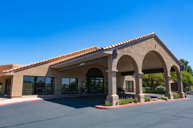Exterior view of La Cañada Care Center building with a covered entrance supported by arches, surrounded by a paved driveway and landscaped with small plants and trees under a clear blue sky.