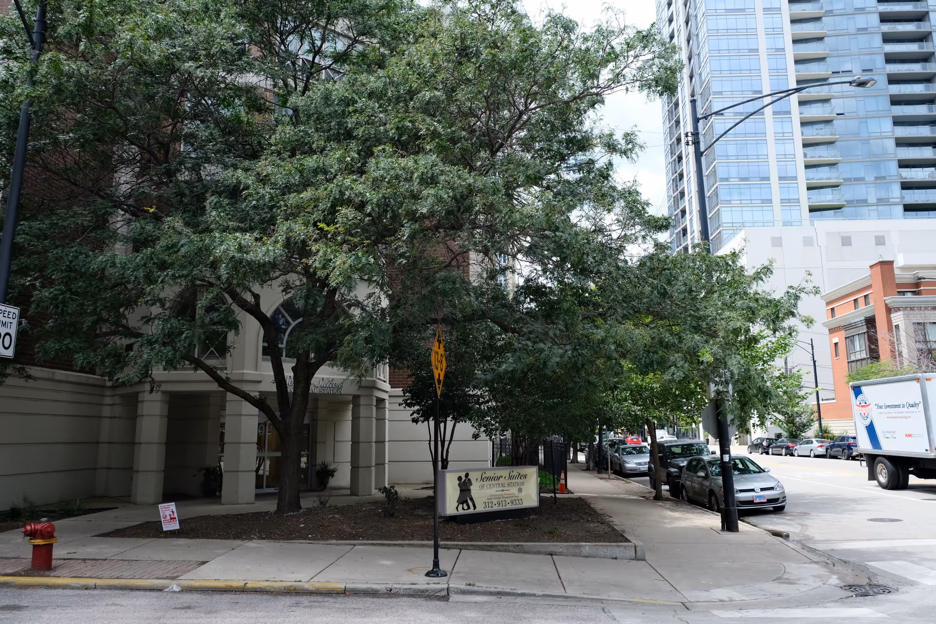 Street-level view of the entrance to a senior living building with large trees, a 'Senior Suites' sign, sidewalk and parked cars in front of nearby high-rises.